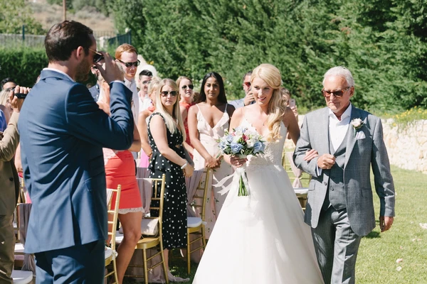 Bride in white with bouquet smiles beside her father at the outdoor wedding ceremony, guests seated