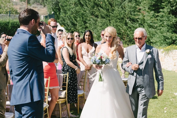 Bride in white with bouquet smiles beside her father at the outdoor wedding ceremony, guests seated