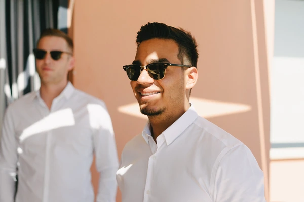 Two boys in white shirts and sunglasses smiling in warm sunshine beside a peach-coloured wall