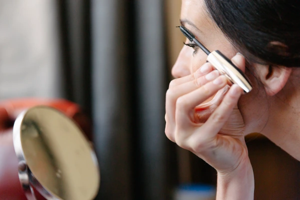 Close-up side profile of the bride applying mascara with a wand, holding a compact mirror, dark hair pinned up. Candid morning preparations.