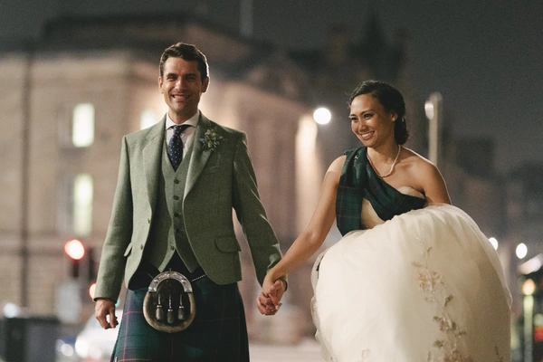 Newlyweds walk hand in hand at night — groom in green jacket and tartan kilt, bride in white lace