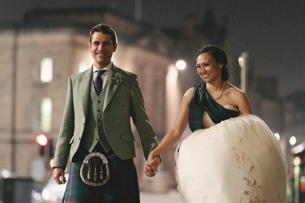 Newlyweds walk hand in hand at night — groom in green jacket and tartan kilt, bride in white lace