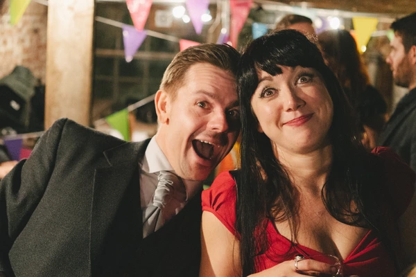 Man in black suit and woman in red dancing amid colourful paper decorations in the lit marquee