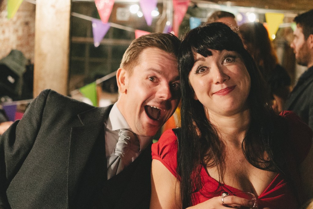 Man in black suit and woman in red dancing amid colourful paper decorations in the lit marquee
