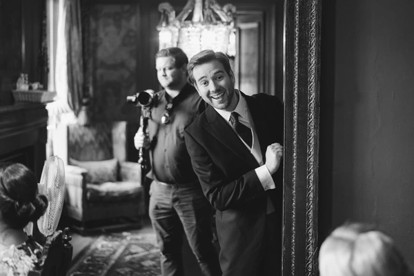 Cheerful groom in a suit laughing and pointing at the camera — black-and-white, crystal chandelier above