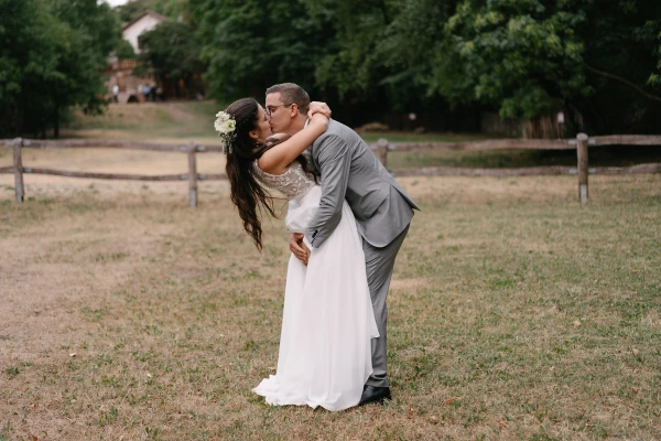 Bride kissing groom at outdoor wedding with white dress and grey suit near wooden fence.