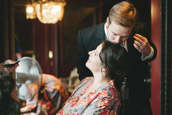 Groom with auburn hair in black suit looks lovingly at the bride in a floral robe