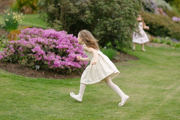 A flower girl in a cream dress runs freely across the garden lawn past pink flowering azaleas at the Royal Botanic Garden Edinburgh. Candid documentary wedding photograph.