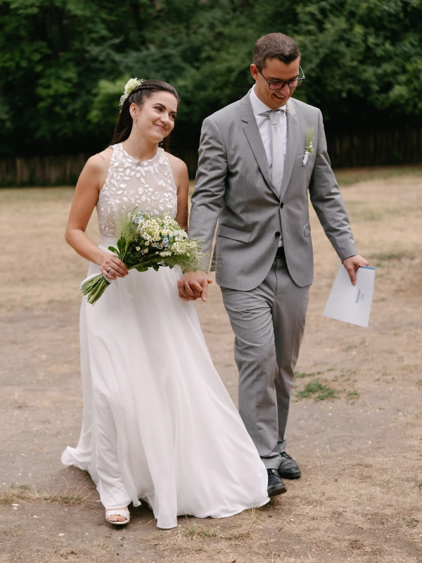 Bride holding white daisy bouquet walking with groom after wedding ceremony outdoor garden venue.