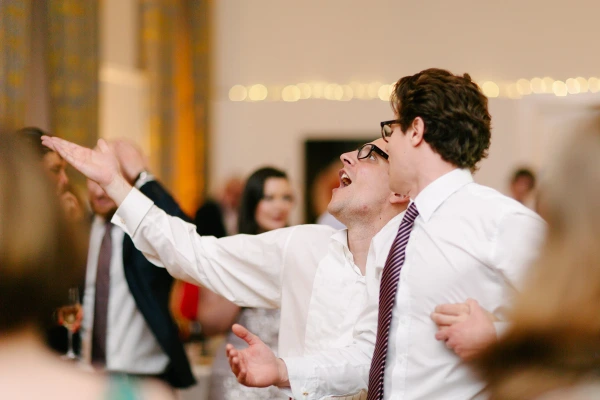 Two women in colourful dresses dance and laugh together on a brightly lit wedding dance floor while a male guest photographs them in the background. Candid documentary wedding photograph.