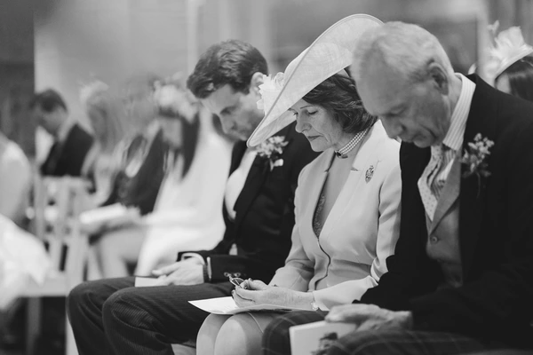 Elegant woman in white hat writes at the ceremony beside a man in a dark suit — focused moment
