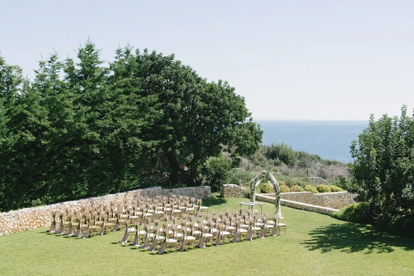 Ceremony venue by the sea — green lawn, antique stone walls, chairs in rows, white floral arch