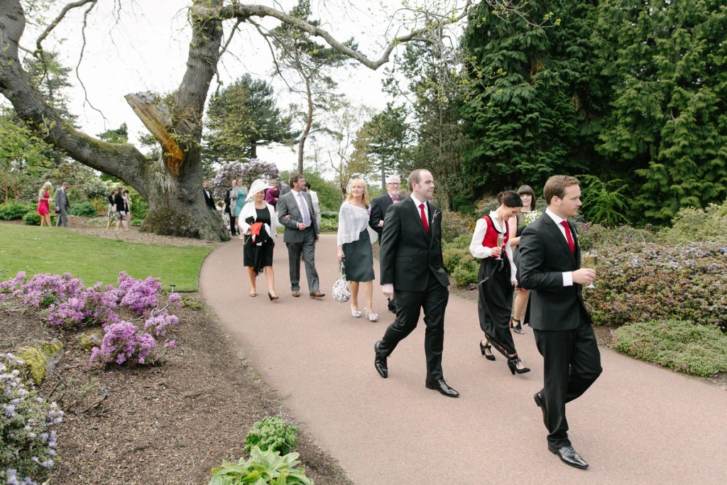 Wedding party and guests walking along a garden path through flowering borders at the Royal Botanic Garden Edinburgh after the ceremony. Candid documentary scene.
