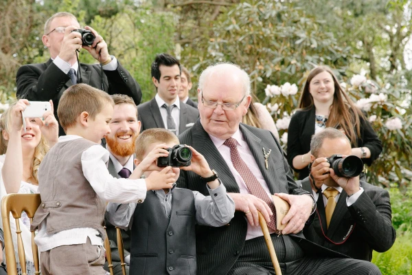 Candid shot of the groom's father with glasses and a red striped bow tie warmly embracing a smiling bridesmaid at the outdoor reception. Joyful post-ceremony candid.