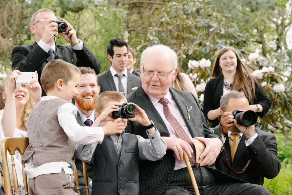 Candid shot of the groom's father with glasses and a red striped bow tie warmly embracing a smiling bridesmaid at the outdoor reception. Joyful post-ceremony candid.