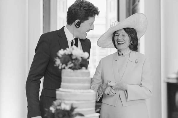 Groom and his mother laughing together at the wedding — white hat, wedding cake and colonnade behind