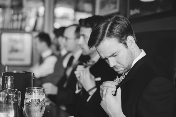 Groom in black suit examines his boutonniere at the table amid glasses at the reception