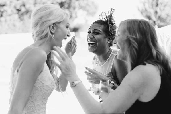 Three women laughing together at the wedding — the middle one has a rose in her hair, holds wine