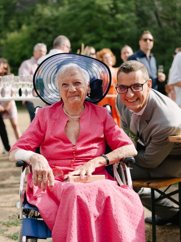 Elderly guest in pink dress and wide-brimmed navy hat posing with groom at outdoor wedding reception.