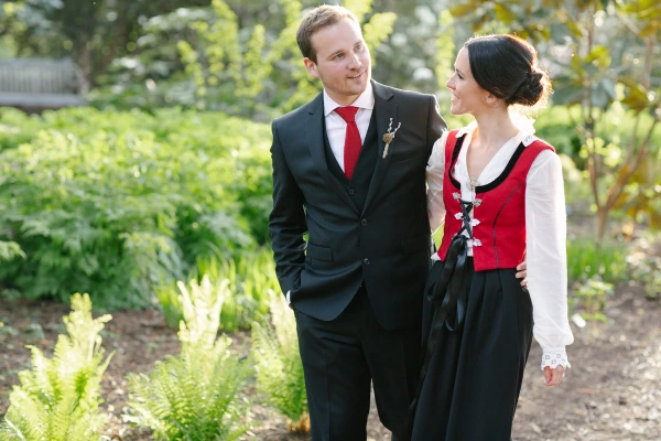 The bride in her Norwegian red bunad and the groom in a dark suit with red tie share a smiling glance while walking through the garden. Candid documentary wedding portrait at the Royal Botanic Garden Edinburgh.
