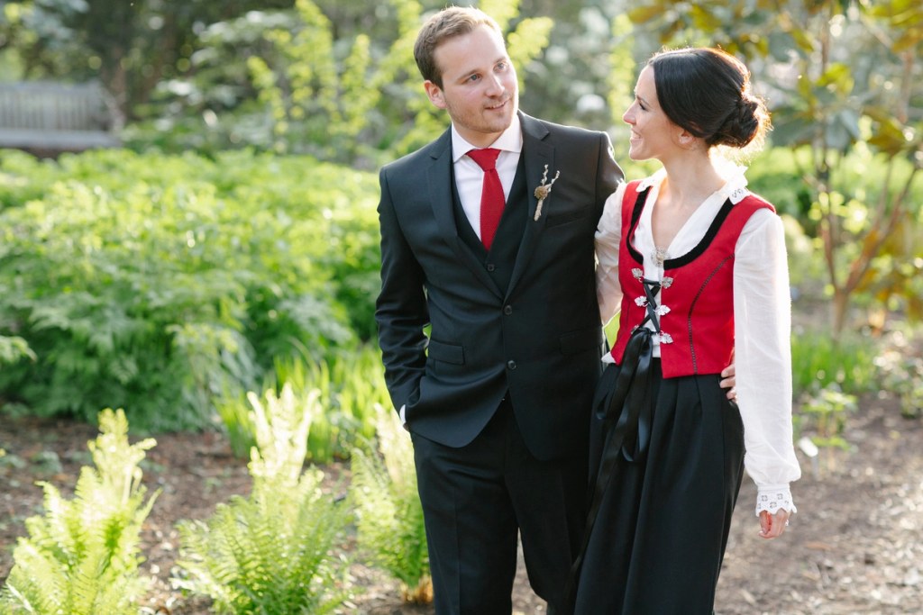 The bride in her Norwegian red bunad and the groom in a dark suit with red tie share a smiling glance while walking through the garden. Candid documentary wedding portrait at the Royal Botanic Garden Edinburgh.