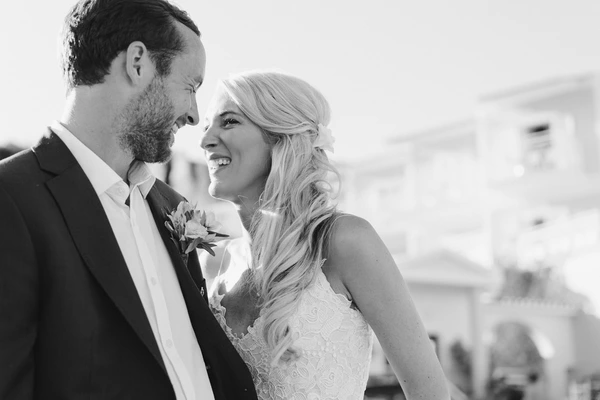 Groom and bride look at each other smiling in sunlight — flower boutonniere, long wavy hair