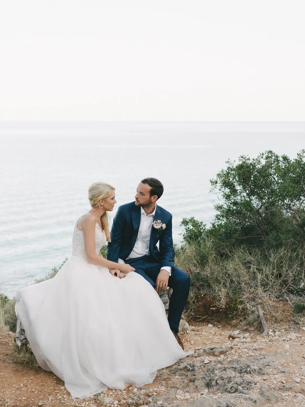 Bride and groom sit hand in hand on a rocky seaside — white gown flowing, clear sky and calm sea