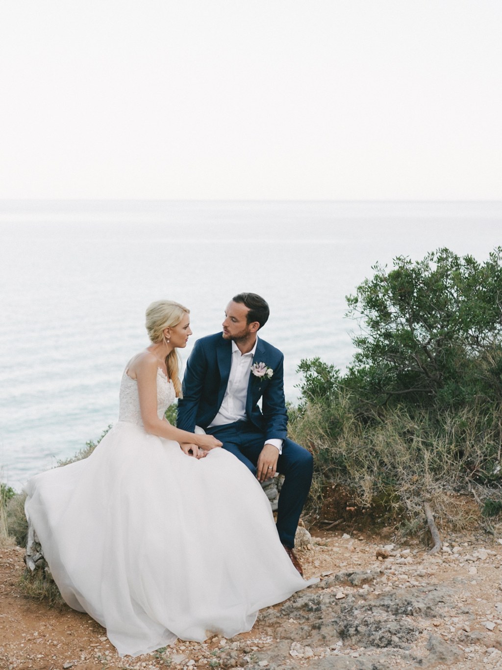 Bride and groom sit hand in hand on a rocky seaside — white gown flowing, clear sky and calm sea