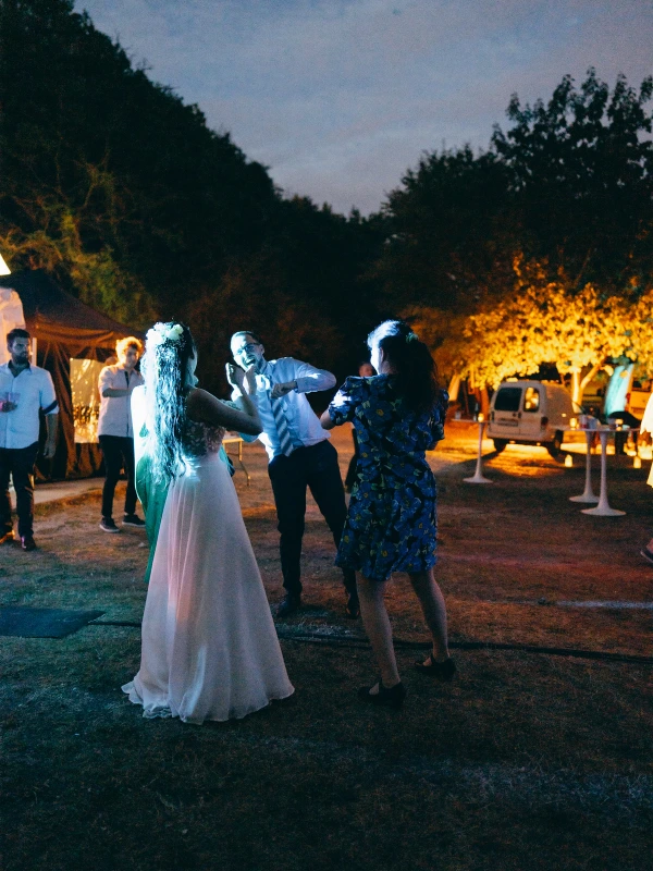 Couple dancing under trees at outdoor evening wedding with warm fire glow and guests.