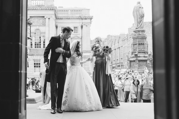 Groom holds bride in his arms on a balcony overlooking a historic square — guests watching