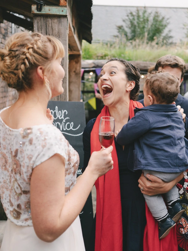 Bride with braided updo talks to a woman in red holding a small boy — joyful wedding moment