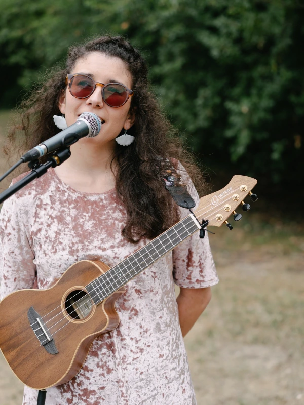 Woman in glasses and a velvet dress singing into a microphone while holding a ukulele. Wedding live music.