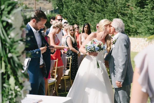 Bride in white with blue-white bouquet stands with father at the altar — guests watch, green garden
