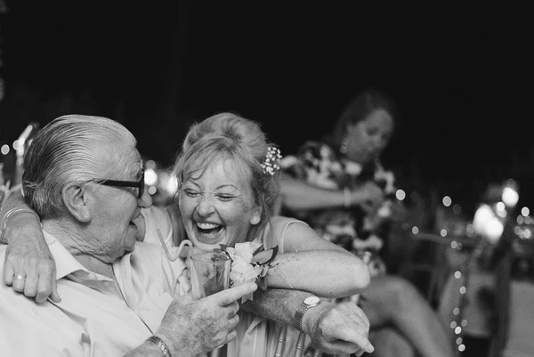 Older man with glasses and woman dance and laugh at the wedding, glass in hand, bokeh lights behind