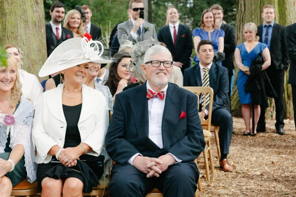 Candid shot of the bride's parents seated in the front row smiling broadly during the outdoor ceremony, wedding guests packed behind them. Joyful family moment.