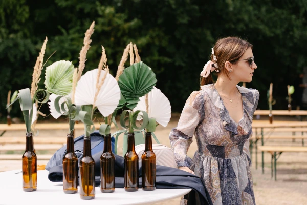 Woman in a floral dress looking away next to beer bottles used as flower vases with paper fans.