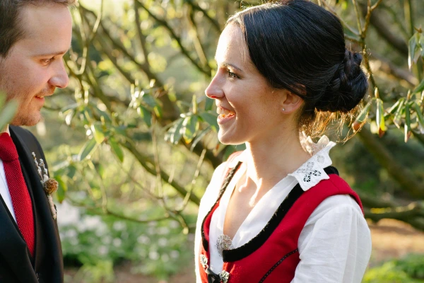 The groom in a dark suit and the bride in a Norwegian red bunad share a warm smile facing each other in the garden's golden evening light. Documentary wedding portrait.