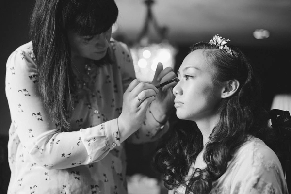 Bride in profile with floral hair detail while makeup artist fixes lash — grey-toned intimate moment