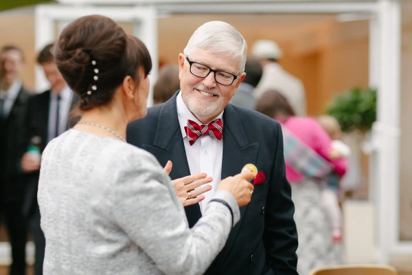 A woman pins a badge onto the lapel of an older man in a black suit with a red striped bow tie at the wedding drinks reception. Candid and warm documentary wedding moment.