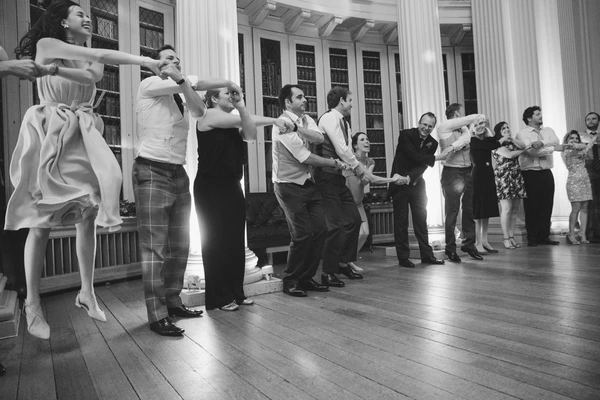 Joyful group dance among wedding guests — white columns and bookshelves, black-and-white
