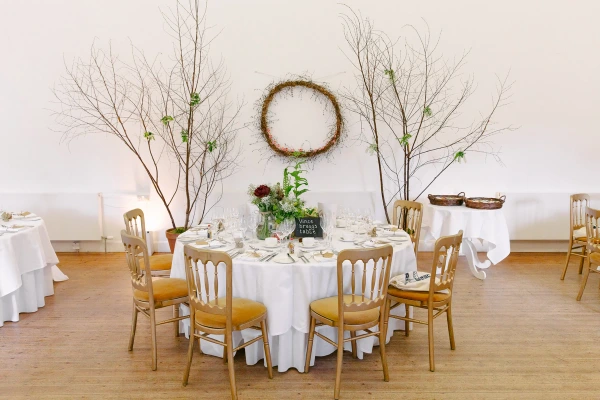 Wide shot of an elegant wedding reception room with round tables set on a wooden floor, bare branch decorations, a wreath on the wall, and golden Chiavari chairs. Venue detail.