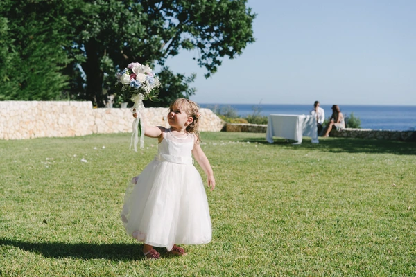 Little girl holds white bouquet up with upward gaze on a green meadow — sea and trees behind