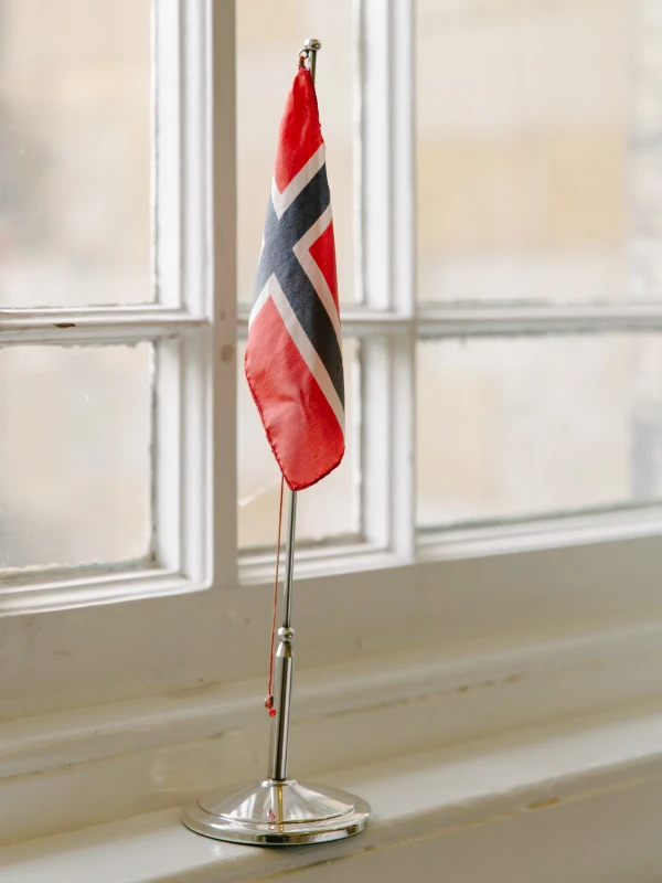 A small Norwegian flag on a chrome stand placed on a window ledge, soft light filtering through the glass. Wedding venue detail.