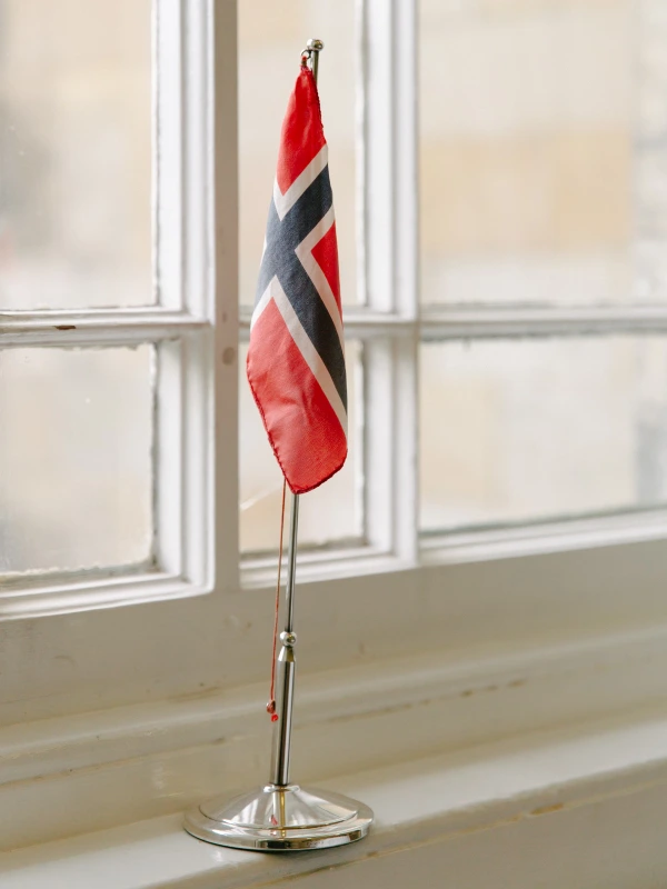 A small Norwegian flag on a chrome stand placed on a window ledge, soft light filtering through the glass. Wedding venue detail.
