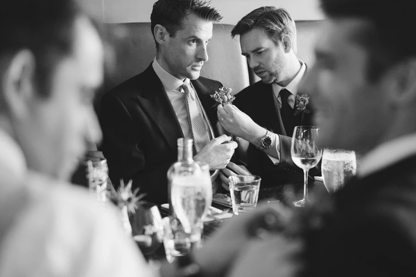 Groom and best man in black suits at the table — floral buttonhole, glasses around them