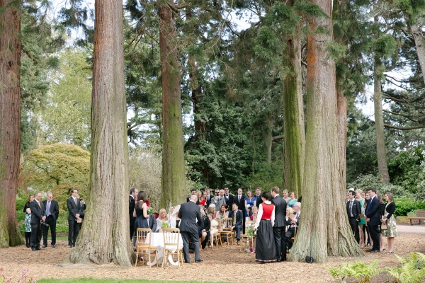 Wide colour shot of the full outdoor ceremony scene among giant redwood trees at the Royal Botanic Garden Edinburgh, guests gathered in a circle around the couple. Documentary overview.