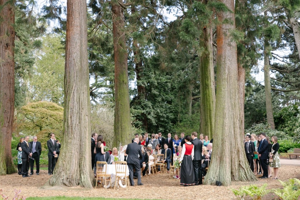 Wide colour shot of the full outdoor ceremony scene among giant redwood trees at the Royal Botanic Garden Edinburgh, guests gathered in a circle around the couple. Documentary overview.