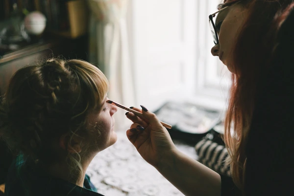 Makeup artist applies brush to bride's face in fresh morning sunlight by the window