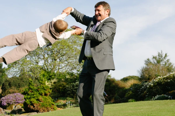 A laughing man in a suit swings a young boy through the air by his arms in the garden of the Royal Botanic Garden Edinburgh. Candid and joyful documentary wedding photograph.