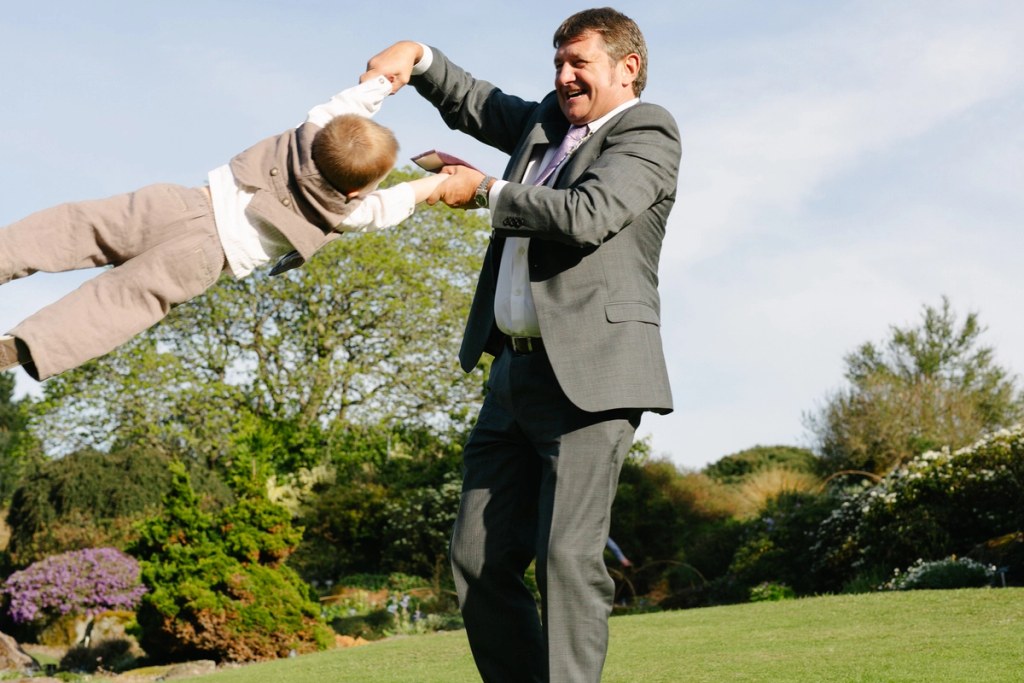 A laughing man in a suit swings a young boy through the air by his arms in the garden of the Royal Botanic Garden Edinburgh. Candid and joyful documentary wedding photograph.